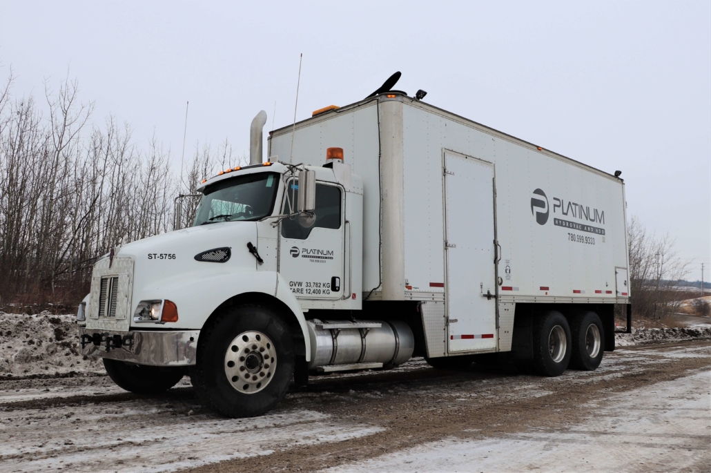 A platinum hydrovac steamtruck parked on the road in front of trees.